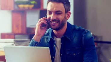 Man in a casual shirt on phone while using laptop, smiling.