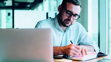 Man writing in notebook at desk with laptop in office setting.