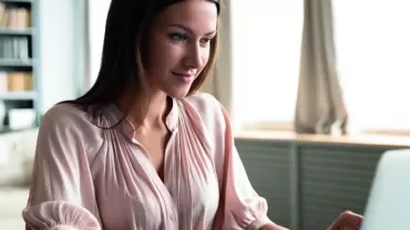 Woman in a pink blouse working on a laptop at home.