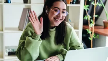 Person in green sweatshirt waving at a laptop during a video call.