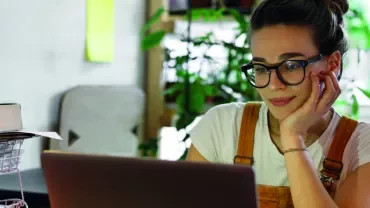 Woman with glasses working on a laptop at a desk with plants in the background.