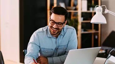 Man smiling while working at a desk with a laptop and lamp.