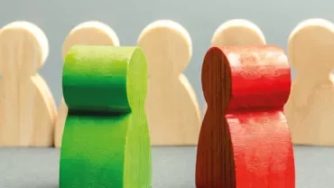 Two wooden figures, one green and one red, with four beige figures in the background.