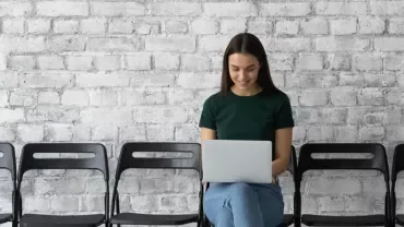 Person sitting with a laptop on a row of chairs against a brick wall.