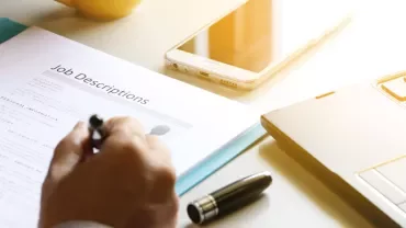 A person writing on a job description beside a laptop and smartphone.