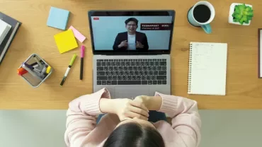 Person watching a video conference on a laptop, with office supplies and coffee on desk.