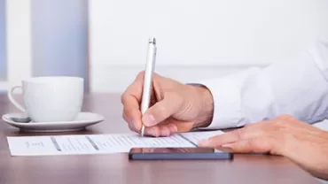Person writing on a document with a cup and smartphone nearby.