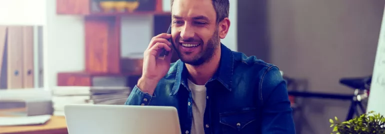 Man in a casual shirt on phone while using laptop, smiling.