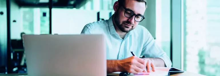 Man writing in notebook at desk with laptop in office setting.