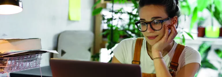 Woman with glasses working on a laptop at a desk with plants in the background.