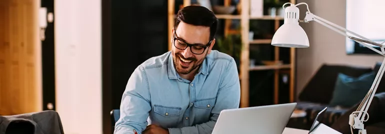 Man smiling while working at a desk with a laptop and lamp.
