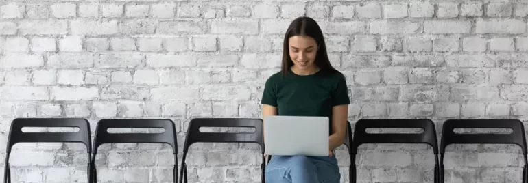 Person sitting with a laptop on a row of chairs against a brick wall.