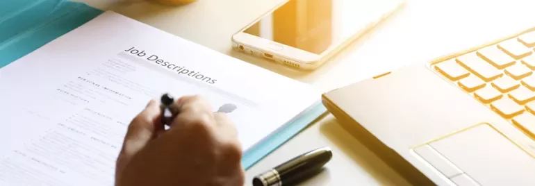 A person writing on a job description beside a laptop and smartphone.