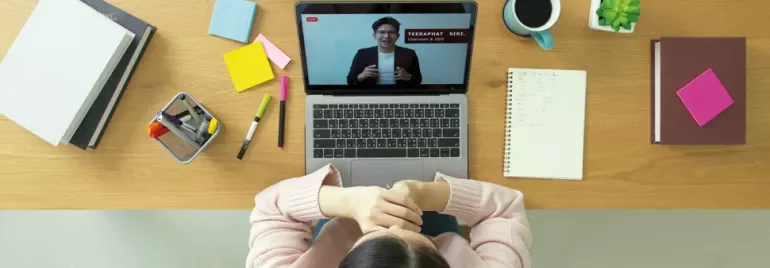 Person watching a video conference on a laptop, with office supplies and coffee on desk.