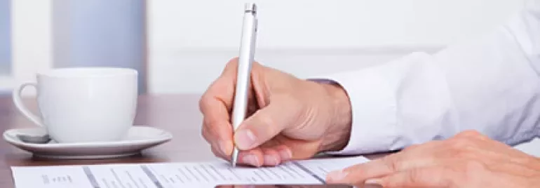Person writing on a document with a cup and smartphone nearby.